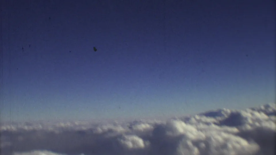 1973 home-movie aerial frame with blue sky dominating the view and cloud banks along the bottom of the aircraft-window scene.