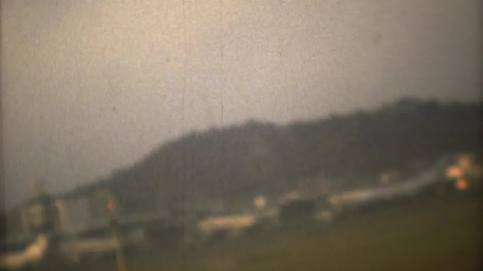 Soft 8mm frame from Mexico City context showing a distant hill, low airport structures, and hazy sky during takeoff.