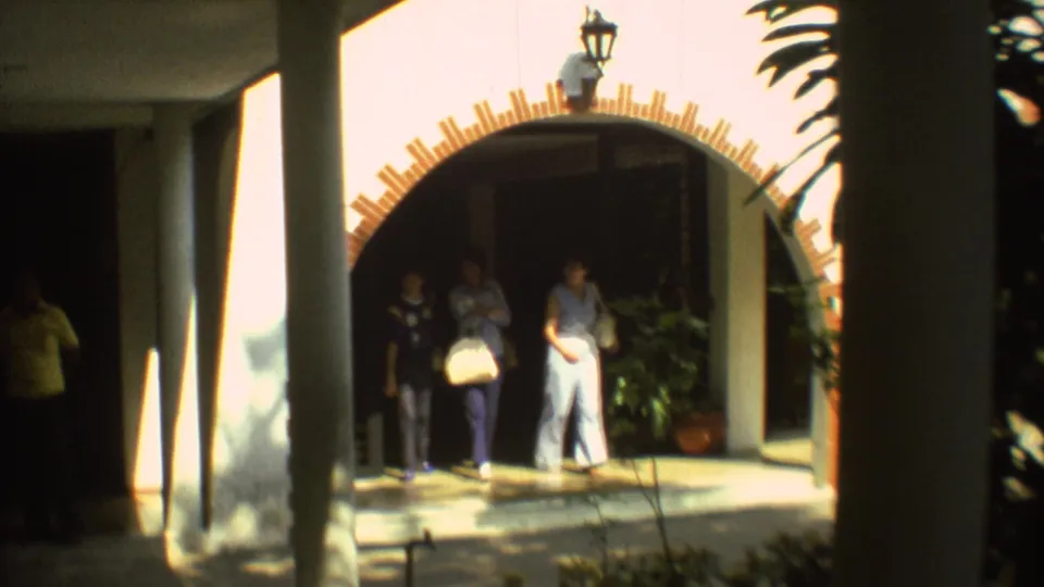 1973 Mexico City frame showing three people at a white building entrance with an arched brick-trim opening and columns.