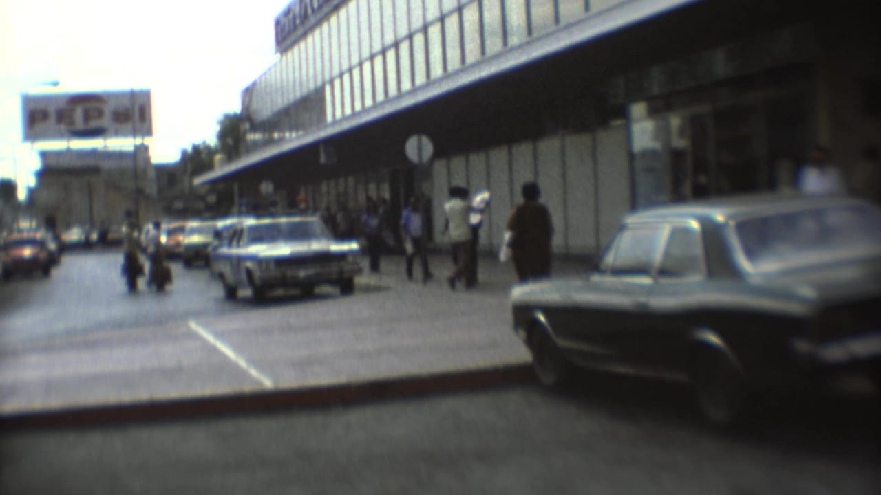 Street with classic cars, pedestrians, a glass-fronted building, and a billboard in the distance.