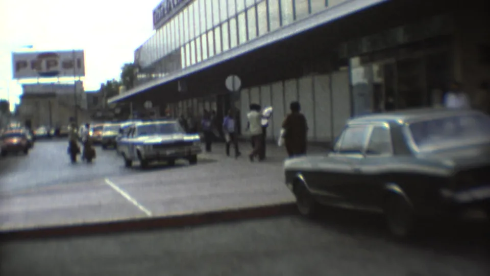 1973 Mexico City street view with older cars, pedestrians on the sidewalk, a modern glass building, and a distant billboard.
