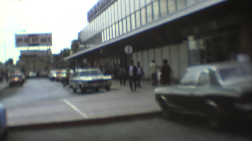 1973 Mexico City street frame with a dark sedan near the camera, pedestrians by a glass building, parked cars, and a billboard.