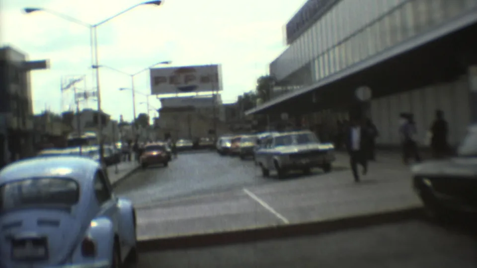 1973 Mexico City street view with a light blue Volkswagen Beetle, moving traffic, pedestrians, streetlights, and a billboard.