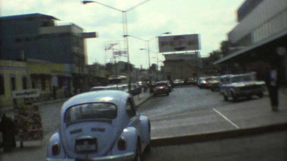 1973 Mexico City traffic scene with a light blue Volkswagen Beetle, classic cars, pedestrians, streetlights, and a visible billboard.