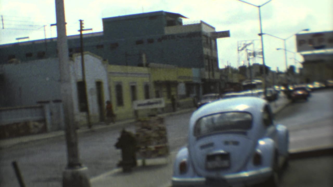 Blue Volkswagen Beetle beside a city street with parked cars, buildings, and pedestrians.