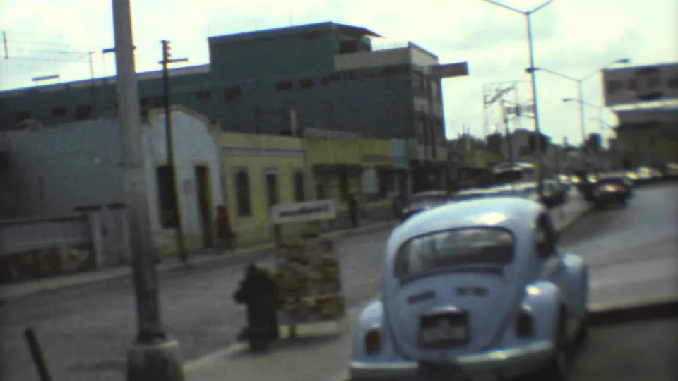 1973 Mexico City street scene showing a blue Volkswagen Beetle, parked cars, pedestrians, and commercial buildings under tall streetlights.