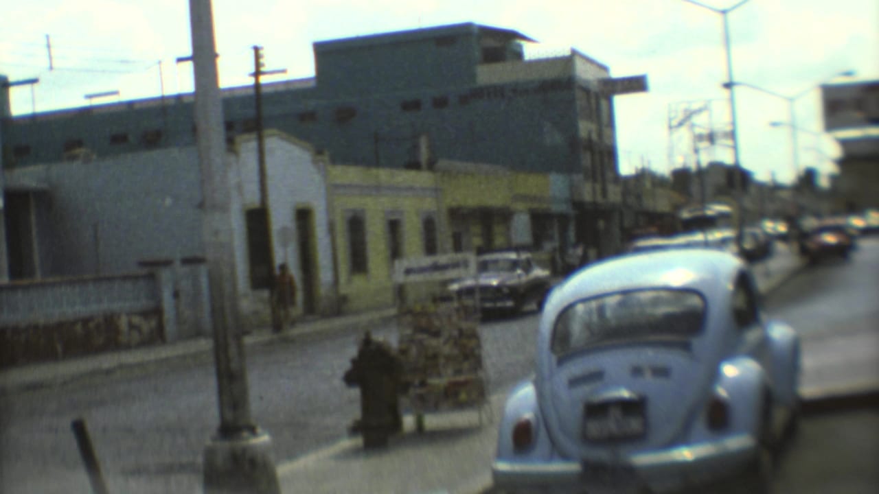 1973 Mexico City street view with a blue Volkswagen Beetle, pedestrians, a small cart or stand, parked cars, and commercial buildings.