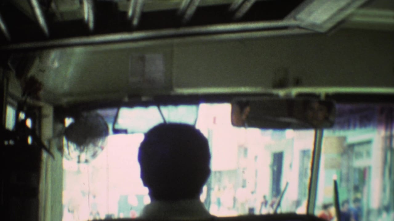Passenger silhouette faces the bus windshield with mirror, fan, and street outside.