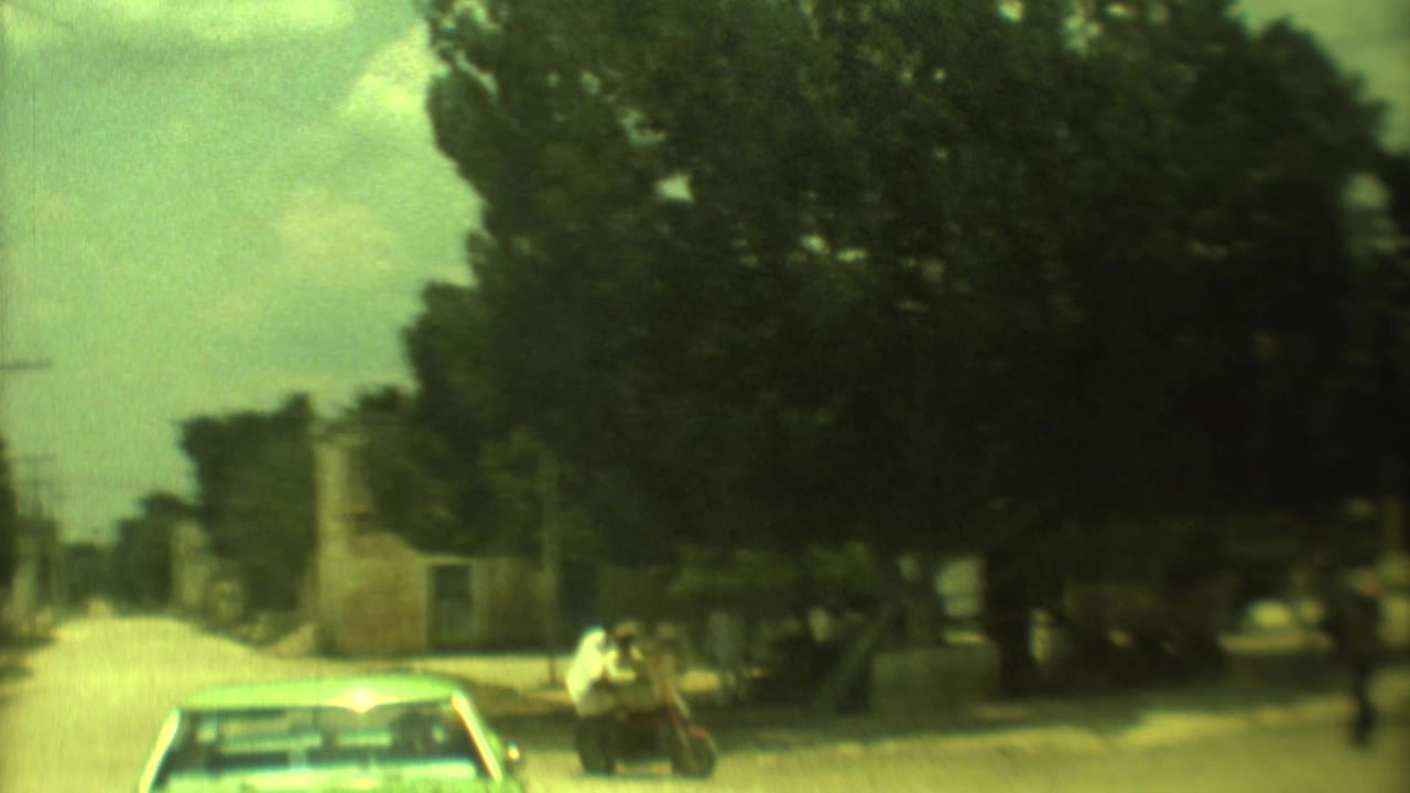 1973 Mexico City home-movie frame showing a shaded roadside, a car, pedestrians, and a motorbike rider on a sunlit street.