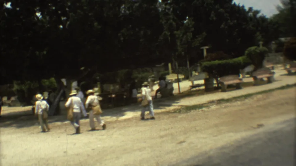1973 Mexico City home-movie frame showing men in light clothing walking through a shaded public area with trees and low structures.