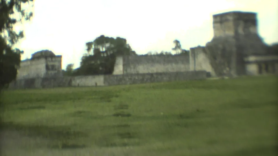 1973 Mexico City 8mm frame showing a wider landmark view across grass, with stone walls and trees in the background.