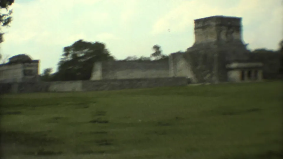 1973 8mm travel frame of stone ruins in Mexico viewed from a grassy area with trees and pale sky.