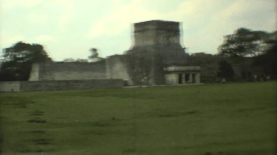 Home-movie view of weathered stone ruins in Mexico, with a stepped wall and trees behind the open grass.
