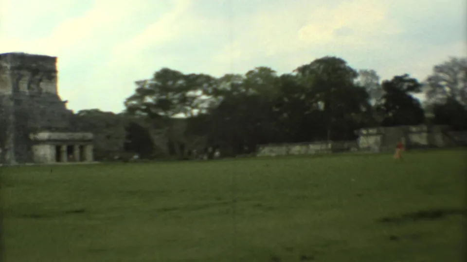 Wide 1973 home-movie frame of Mexican stone ruins with lawn, trees, and tiny distant visitors at the site.