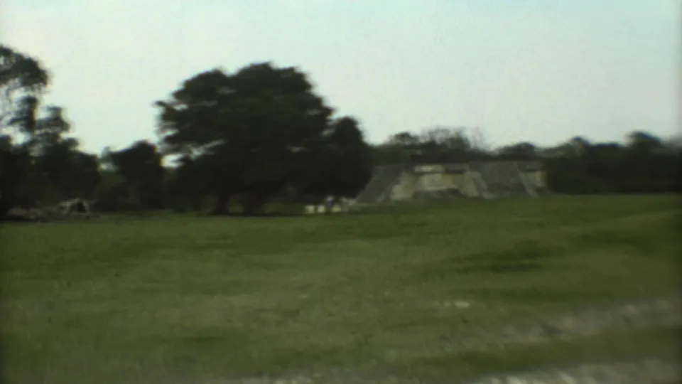 1973 Mexico City home-movie frame with an open field, tree silhouettes, and a distant stone ruin or wall at the edge of view.
