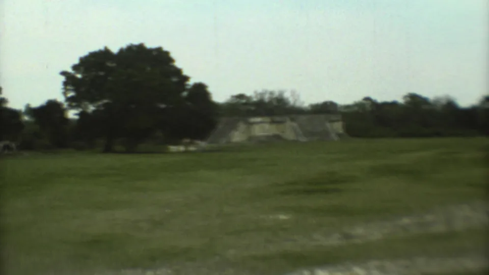 1973 8mm travel footage shows a distant stepped temple across grass at a Mexican archaeological site.