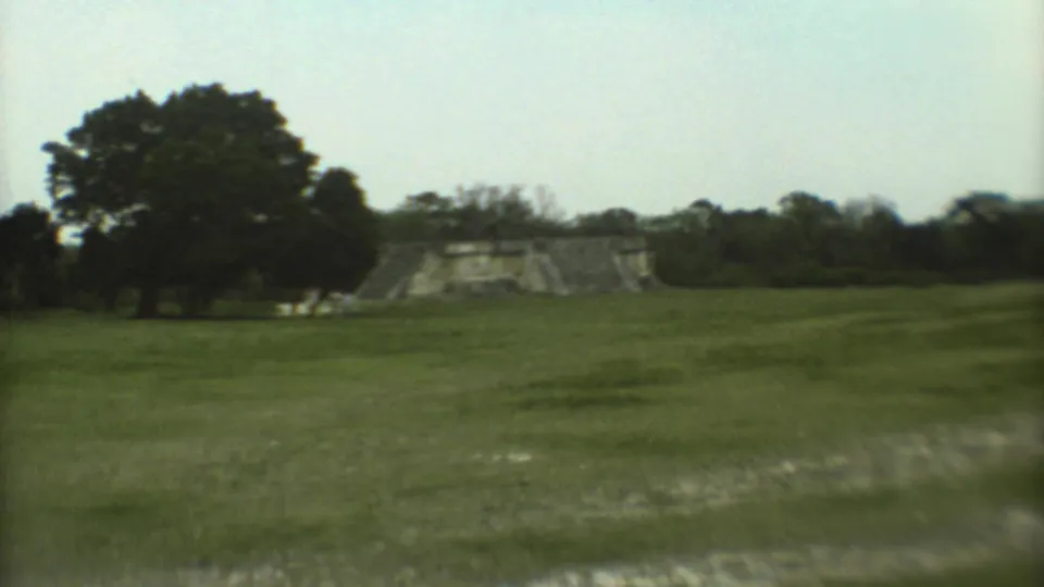 The camera holds on the temple from a distance, with trees and open lawn dominating the frame.