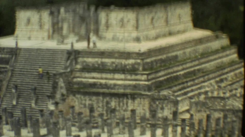 Wide 8mm frame of ancient stone steps and platforms in Mexico City, with visitors scattered across the ruin in the distance.