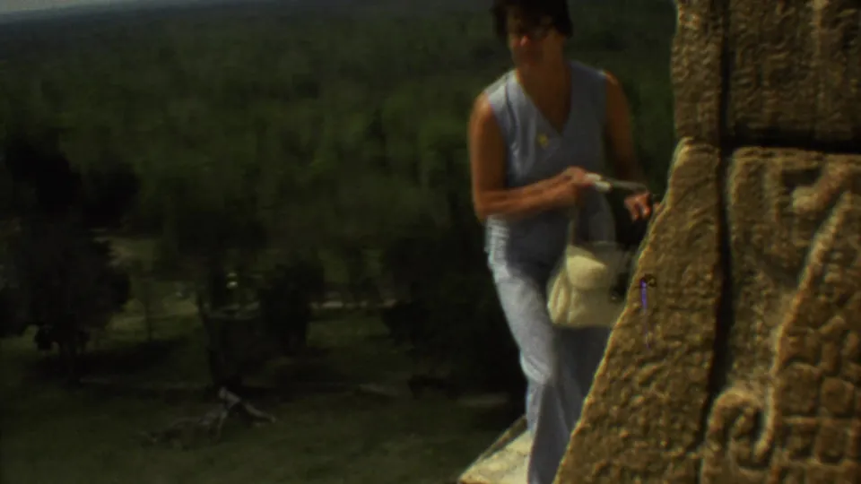 A woman in pale blue clothing stands near carved stone masonry at the ruins, with dark green landscape visible beyond her.