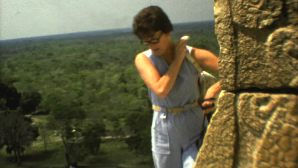 Close 1973 travel-film view of a woman leaning near carved stone blocks, with forested land stretching behind the ruin.