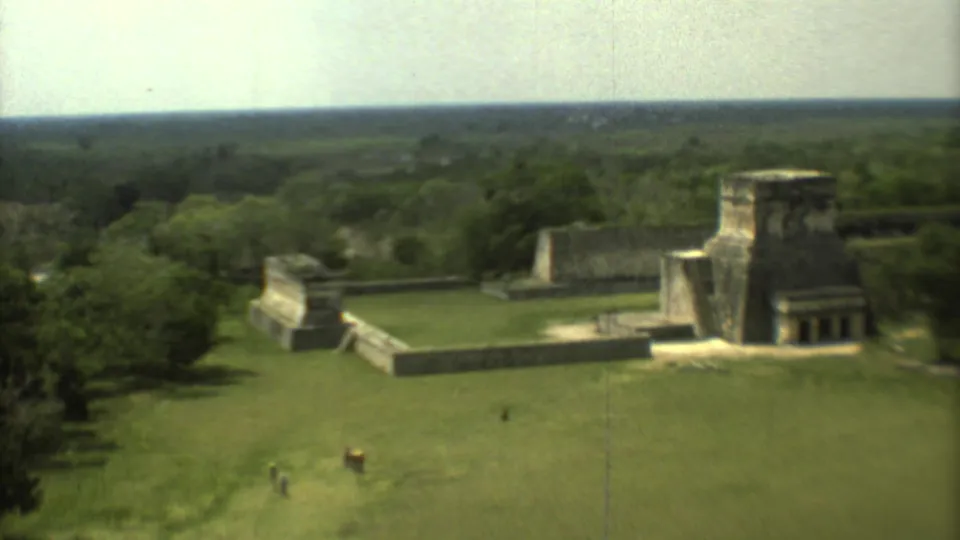 1973 8mm aerial view of a Mesoamerican ruin complex in Mexico, with stone structures, grass, trees, and tiny visitors below.