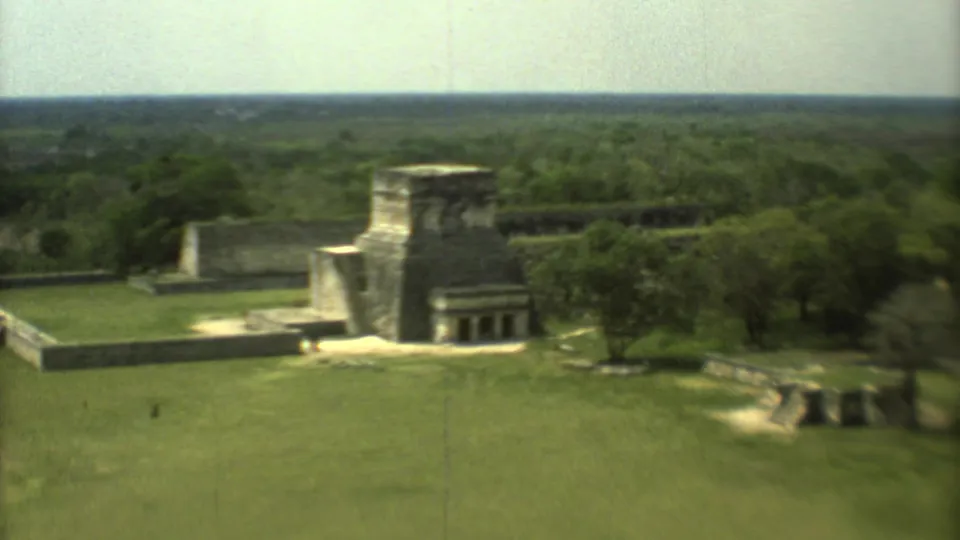 Soft 8mm aerial frame of 1973 Mexico ruins, showing stone architecture, green grounds, paths, trees, and distant visitors.