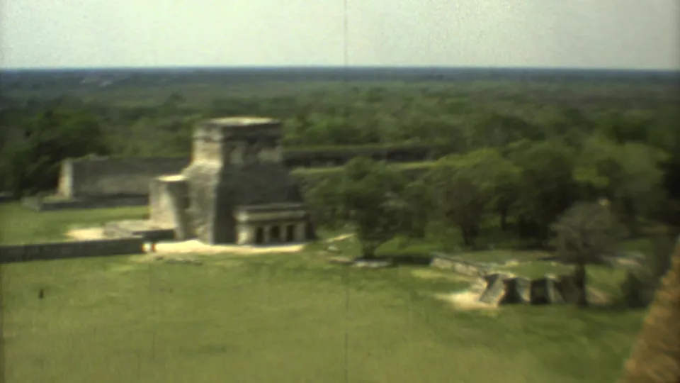 1973 8mm view in Mexico showing stone ruins across a grassy archaeological site with dense trees on the horizon.