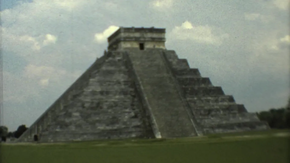 1973 8mm frame of a large stepped Mesoamerican pyramid in Mexico, with grass below and clouds overhead.