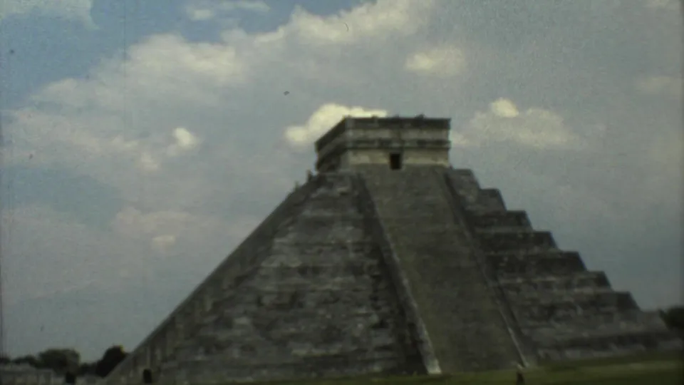 Wider 1973 home-movie frame of the stone pyramid and summit temple, with the broad stairway centered below a cloudy sky.