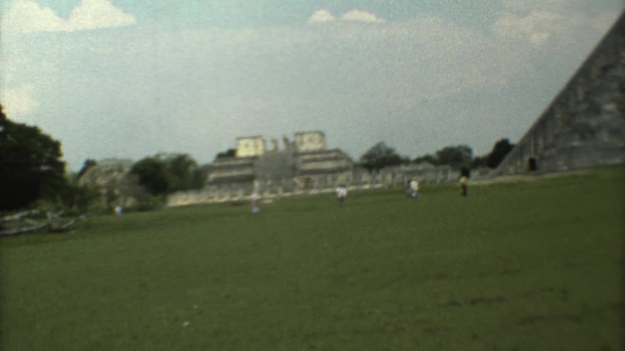 1973 8mm visitor-shot frame showing people scattered across a green field near ruins and the pyramid at the archaeological site.