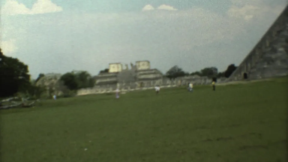 1973 8mm visitor-shot frame showing people scattered across a green field near ruins and the pyramid at the archaeological site.