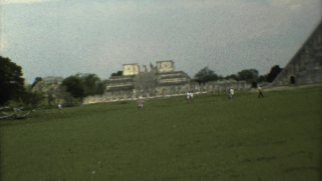Wide grassy field with distant stone ruins, trees, and small visitors under a pale sky.