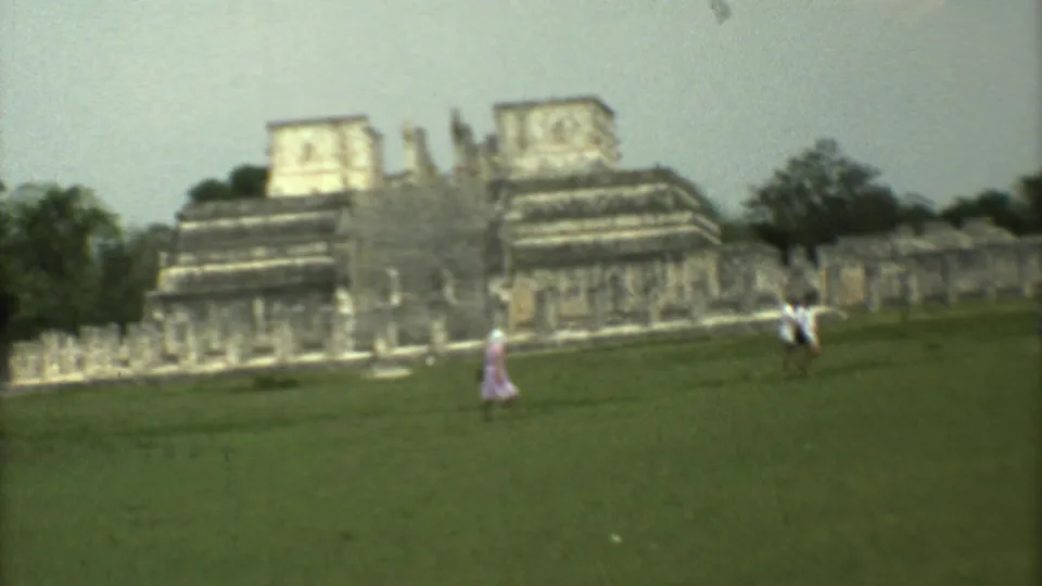 Visitors cross the grass before large stone ruins in Mexico City, including a person in pink near the center foreground.