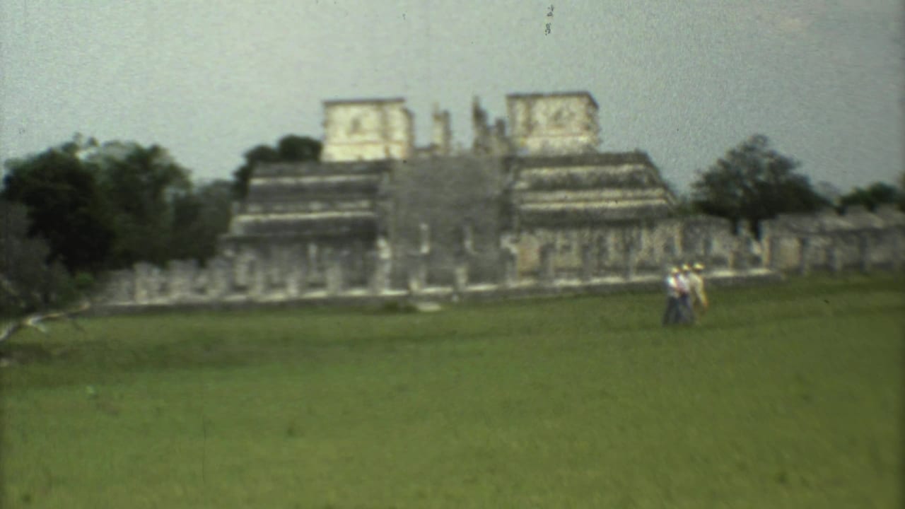 1973 8mm travel frame of tourists on a grassy field before stepped stone ruins in Mexico, with trees and pale sky behind.
