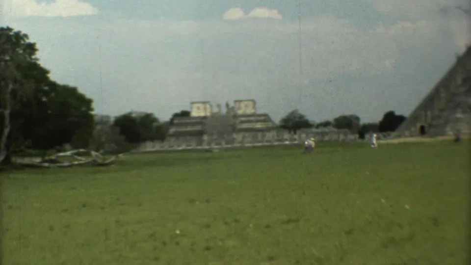 1973 Mexico travel footage frame showing tourists on a lawn near stepped ruins, trees and fallen branches in the foreground.