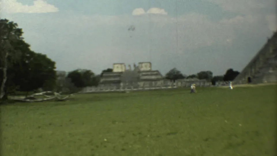 Final 1973 8mm frame of a grassy archaeological site in Mexico with distant tourists, stepped ruins and a pyramid side under clouds.
