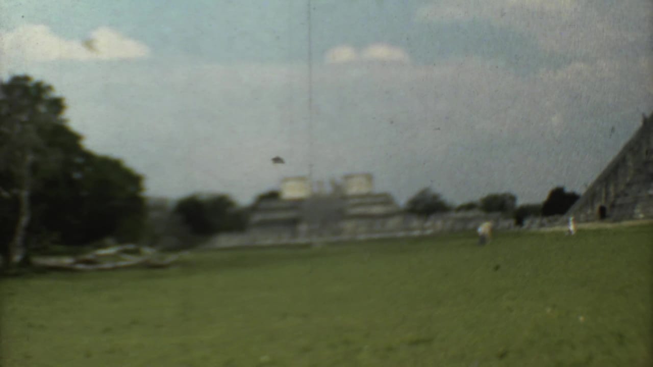 Wide grassy field facing a stepped stone pyramid under a cloudy sky.