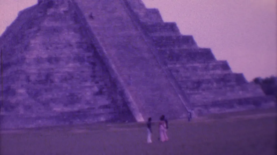 1973 8mm view in Mexico City of tourists at the grassy base of a large stepped temple under a hazy purple sky.