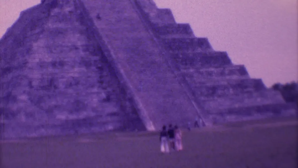 Tourists in 1970s travel clothing gather at the base of a large stone pyramid in Mexico City, seen through faded 8mm color.