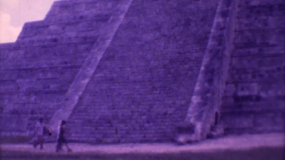 Two tourists walk across the foreground below the broad steps of a large temple structure in 1973 Mexico City home-movie footage.