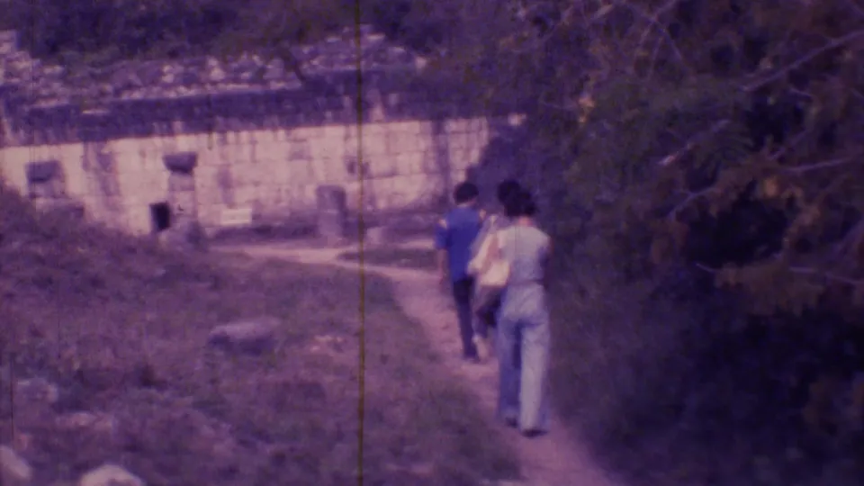 Two visitors seen from behind on a path near greenery and a low historic-looking structure in 1973 Mexico City footage.