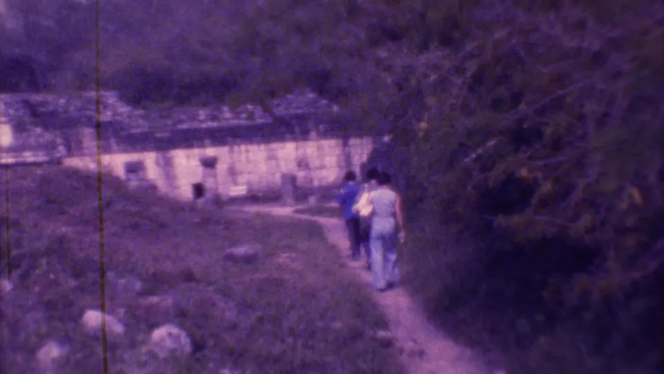 Distant rear view of visitors walking near dense vegetation and a weathered structure in Mexico City archival home-movie footage.