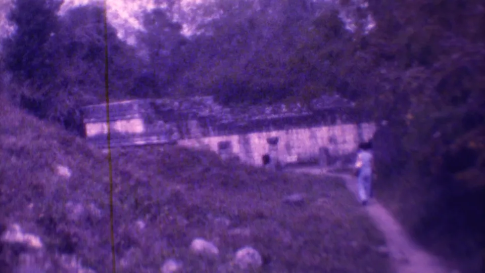 A person in light clothing walks on a narrow path near a low building in wooded 1973 Mexico City footage.