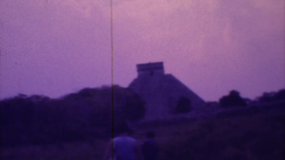 Handheld 1973 Mexico City travel frame with visitors in the foreground and a pyramid-like ruin across hilly, vegetation-covered ground.