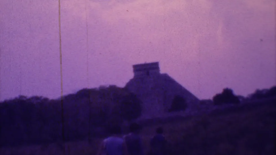 Purple-tinted 8mm frame from 1973 Mexico City showing visitors in open terrain and a distant stepped archaeological structure.