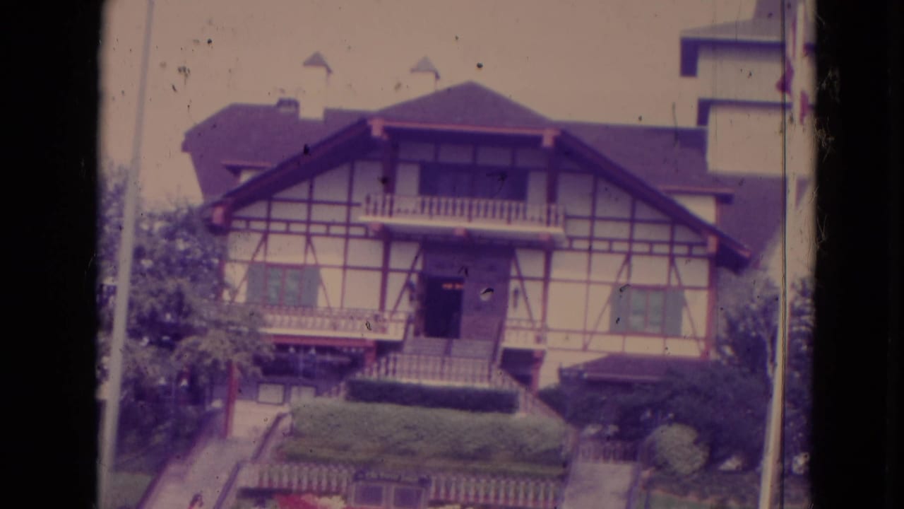 Swiss-style house with timber framing, balcony, shutters, hedges, and garden beds.
