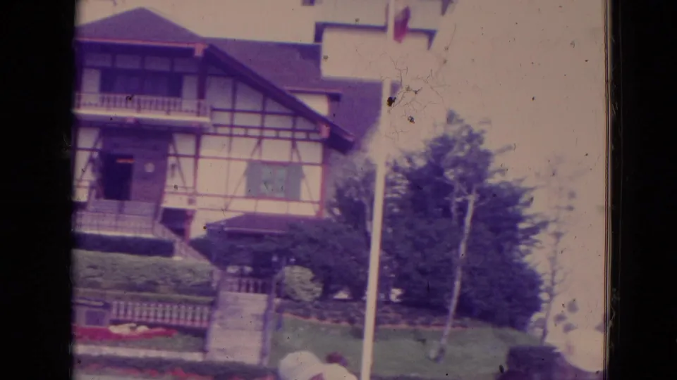 1974 Orlando frame with the Swiss-style house partly centered, showing front steps, hedge rows, lawn, flowerbeds, and a flagpole.