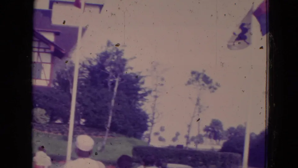 1974 Orlando 8mm frame during the pan, with the house partly cropped, a flagpole, trees, hedges, lawn, and distant visitors.