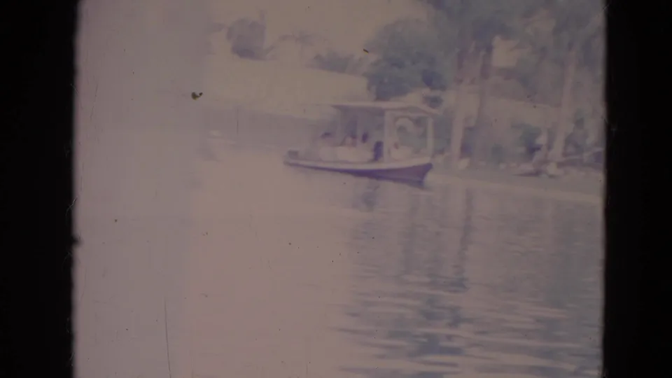 1974 Orlando 8mm frame showing a small covered boat on pond water, with trees, reflections, and distant people visible.