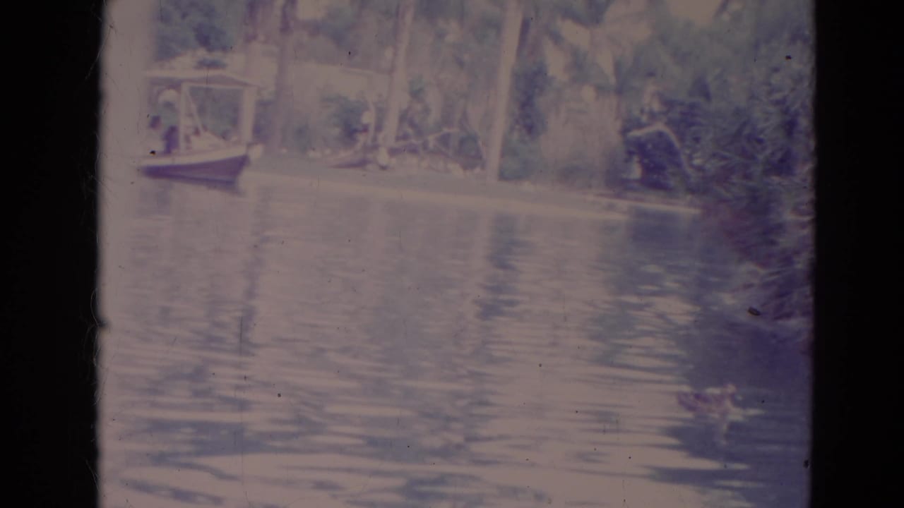 1974 Orlando home-movie frame of a covered boat by a landscaped pond edge, with distant people and dense foliage beyond.